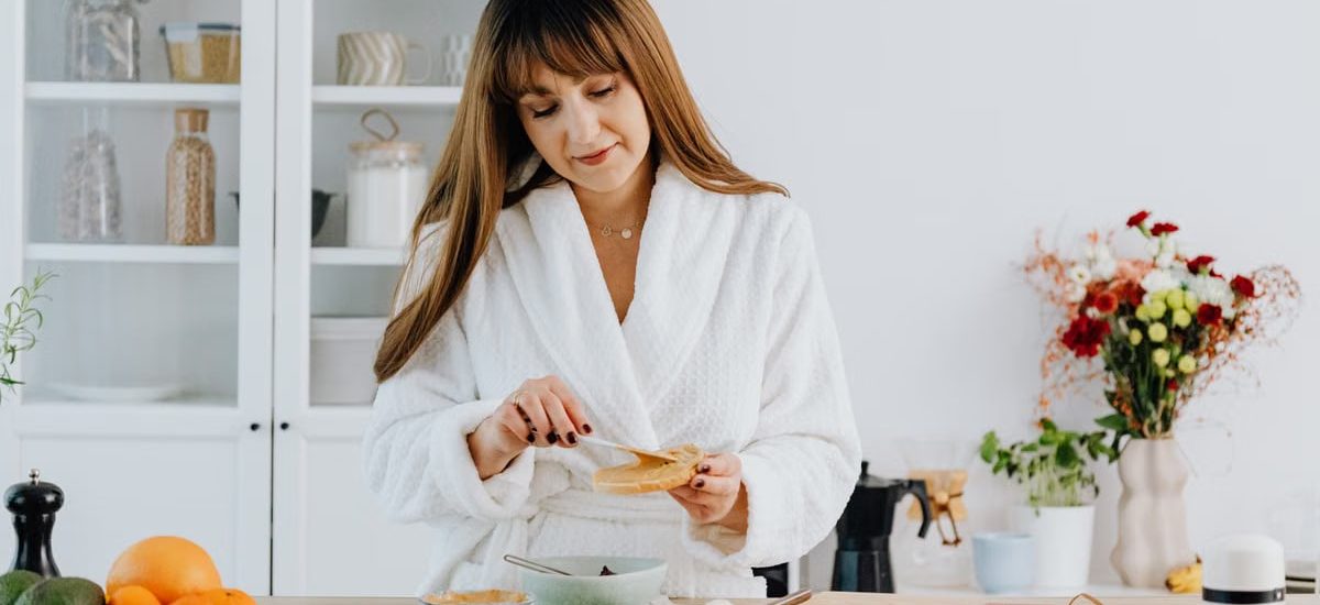A woman in a bathrobe preparing breakfast in a home kitchen.