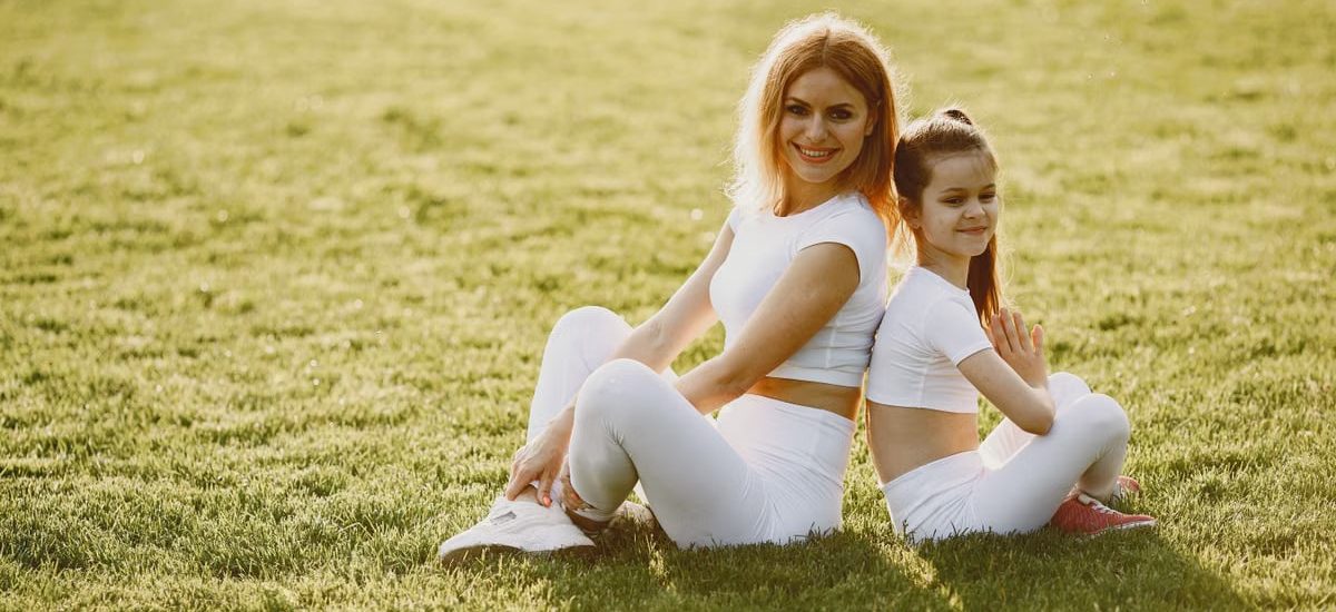A mother and daughter sit back to back in matching activewear on a sunny grass field, enjoying a summer day together.