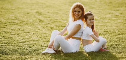 A mother and daughter sit back to back in matching activewear on a sunny grass field, enjoying a summer day together.