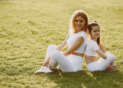 A mother and daughter sit back to back in matching activewear on a sunny grass field, enjoying a summer day together.